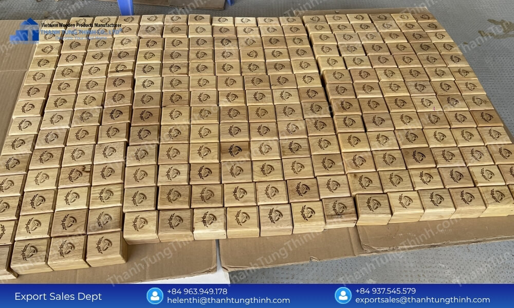 Hundreds of small square wooden boxes with uniform laser-engraved ring logos laid out neatly across cardboard sheets on the floor