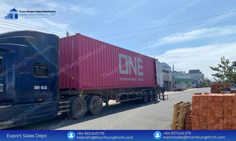 A large dark blue commercial transport truck carrying a long bright pink ONE shipping container parked next to a stack of orange bricks